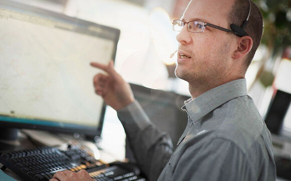 Hotline Ein Mann mit Brille und Headset sitzt an einem Schreibtisch vor einem Computerbildschirm, der eine Karte zeigt. Er zeigt mit dem Finger auf die Karte, während er die Tastatur bedient.