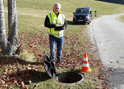 Ein Mann in Warnweste steht am Straßenrand neben einem offenen Kanalschacht; daneben liegt der Deckel, und ein orangefarbener Verkehrskegel steht. Im Hintergrund rollt ein Auto vorbei, Herbstlaub liegt im Gras.