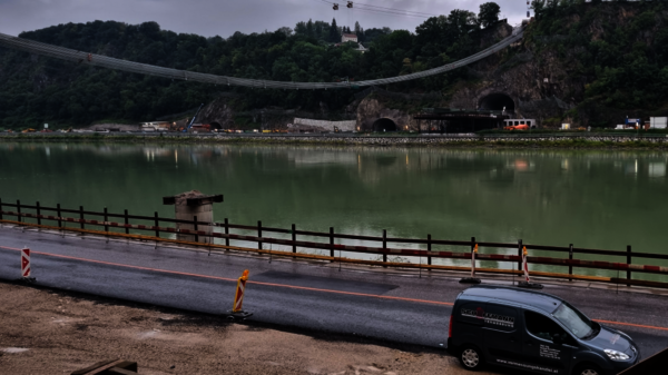 Suspension bridge crosses a calm river framed by forested hills. In the foreground, a paved road with a barrier runs along the water, and a van is parked nearby; a tunnel entrance sits in the rocky hillside behind the bridge.