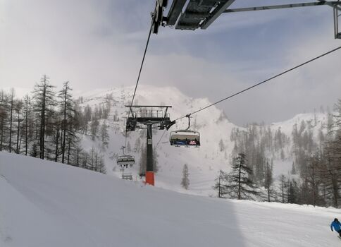 Ski-Lift mit aufgehängten Sesseln zieht über eine verschneite Piste durch eine winterliche Gebirgslandschaft. Im Hintergrund erstrecken sich schneebedeckte Berge und Nadelbäume, während am unteren rechten Rand eine Person in blauem Wintersportoutfit zu sehen ist.