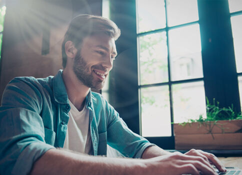Ein lächelnder Mann sitzt an einem Laptop an einem hellen Arbeitsplatz, beleuchtet von Sonnenlicht durch große Fenster im Hintergrund.