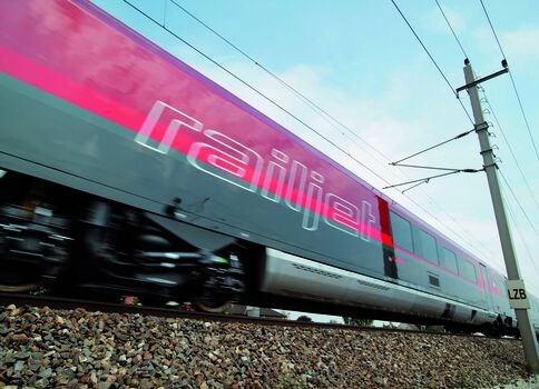 Ein farbenfroher Hochgeschwindigkeitszug mit der Aufschrift Railjet fährt schnell vorbei, rosa und türkis lackiert, auf Schottergleisen; darüber Oberleitungen und ein Mast im Hintergrund.