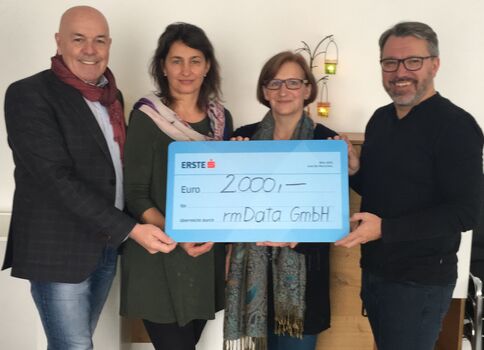 Vier Erwachsene stehen nebeneinander und halten gemeinsam einen großen blauen Scheck. Darauf steht der Betrag 2.000 Euro mit dem Empfänger rmData GmbH; alle lächeln bei der Scheckübergabe.