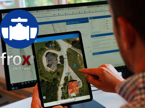 Eine Person nutzt ein Tablet, das eine Luftbildkarte eines Wohnviertels zeigt. Die Hand tippt auf das Display; im Hintergrund sieht man einen Laptop mit einer Dashboard-Oberfläche.