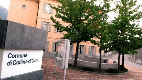 Straßenansicht: Weißes Schild mit der Aufschrift "Comune di Collina d'Oro" vor einem hellorange verputzten Gebäudekomplex. Bäume rahmen die Szene.