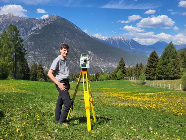 A man stands beside a bright yellow tripod-mounted surveying instrument in a grassy alpine field, with mountains, trees, and a blue sky with scattered clouds in the background.