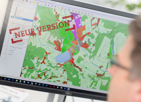 Monitor zeigt eine geografische Karte in GIS-Software mit grünen Feldern und bunten Bereichen; ein roter Stempel "NEUE VERSION" liegt darüber.