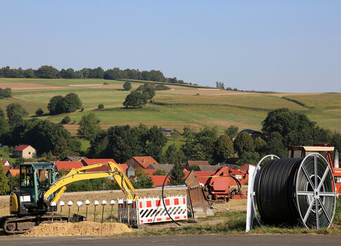 Auf einer ländlichen Straße finden Bauarbeiten statt; ein gelber Bagger gräbt neben Absperrungen, rechts liegt eine große Kabelspule. Im Hintergrund erstrecken sich Felder, Bäume und eine kleine Siedlung mit roten Dächern.