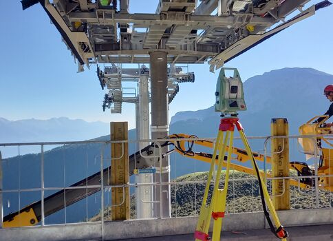 Seilbahnturm in den Bergen mit Wartungsarbeiten: Zwei Techniker in Schutzhelmen arbeiten auf einer Plattform, Geländer sichern den Rand; im Hintergrund erstrecken sich Bergkette und blauer Himmel.