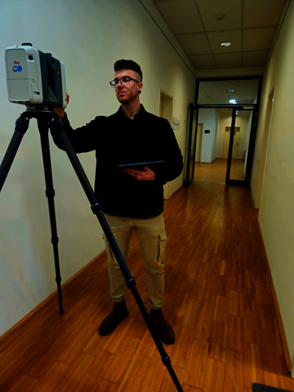 Ein junger Mann mit Brille steht in einem hellen Flur und richtet ein Vermessungsgerät auf einem Stativ aus; er hält ein Tablet in der anderen Hand.