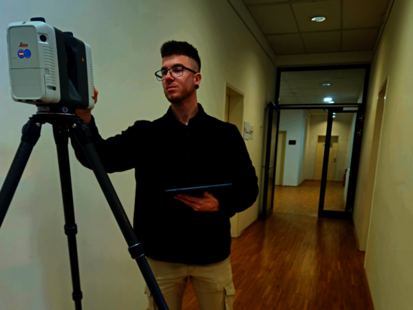 Ein junger Mann mit Brille steht in einem hellen Flur und richtet ein Vermessungsgerät auf einem Stativ aus; er hält ein Tablet in der anderen Hand.