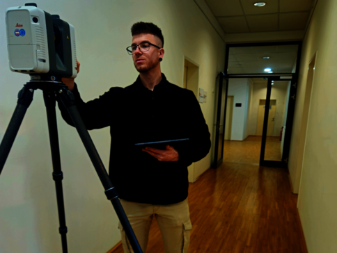 Ein junger Mann mit Brille steht in einem hellen Flur und richtet ein Vermessungsgerät auf einem Stativ aus; er hält ein Tablet in der anderen Hand.