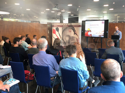 Eine Gruppe Menschen sitzt in blauen Stühlen in einem Konferenzraum und hört einem Redner am Rednerpult zu, der eine Präsentation vor einer Bildschirmprojektion zeigt.