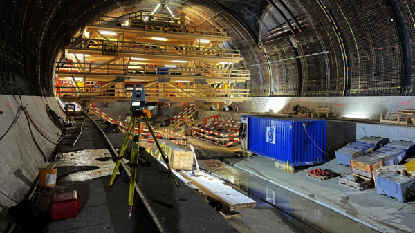 Baustelle in einem Tunnel: Eine mehrstöckige Holzgerüstkonstruktion ragt aus dem Boden; vorne steht ein Vermessungs-Dreibein. Absperrband, Bauwerkzeuge und Baumaterialien liegen herum. Rechts eine blaue Containerkiste, links Paletten; auf dem Boden sind Pfützen sichtbar.