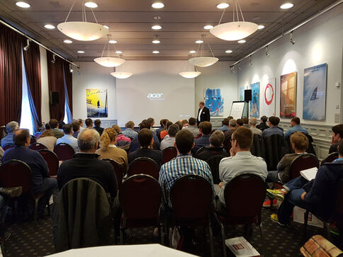 rmDATA Geomatik Event 2019 Viele Zuhörer in roten Stühlen sitzen in einem hellen Saal und hören einem Redner am Podium zu; hinter ihm erscheint eine Projektionsfläche mit dem Acer-Logo.