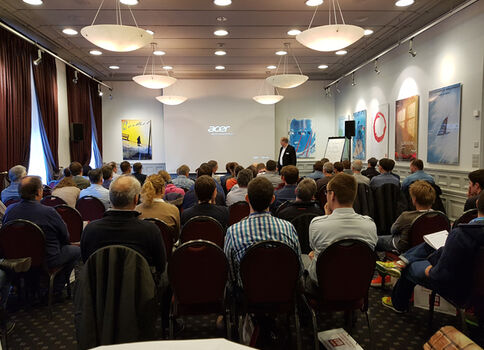 Viele Zuhörer in roten Stühlen sitzen in einem hellen Saal und hören einem Redner am Podium zu; hinter ihm erscheint eine Projektionsfläche mit dem Acer-Logo.