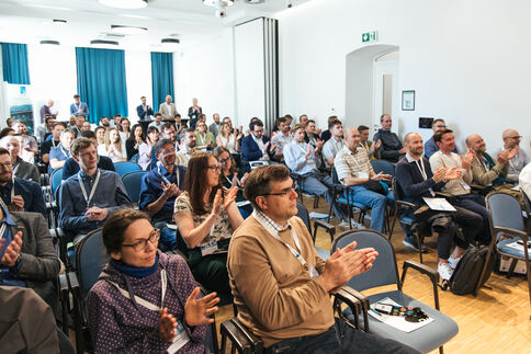 Voll besetzter Konferenzsaal mit vielen lächelnden Zuschauern; einige klatschen, andere fotografieren oder notieren, während eine Präsentation läuft. Der helle Raum hat blaue Vorhänge, weiße Wände und Holzfußboden.