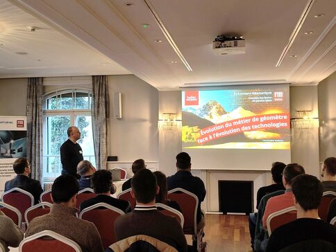 Ein Referent präsentiert eine Präsentation vor einem Publikum in einem Konferenzraum. Die Folie behandelt die Entwicklung des Berufs Geometer im Kontext technologischer Veränderungen.