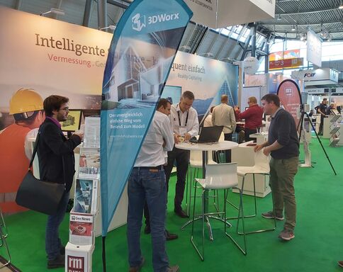 Auf einer Messe steht eine Gruppe Fachleute an einem hohen Tisch mit Laptop, vor einem Ausstellerstand von 3D Worx. Hinter ihnen zeigen Banner Werbung für intelligente Vermessung; weitere Messestände und Besucher sind im Hintergrund zu sehen.