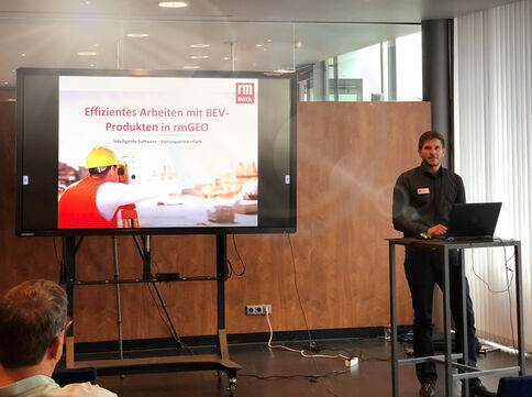 Präsentation in einem Besprechungsraum: Ein Mann steht am Rednerpult mit Laptop; auf dem großen Bildschirm ist eine Folie mit dem Titel „Effizientes Arbeiten mit BEV-Produkten in rmGEO“ zu sehen.