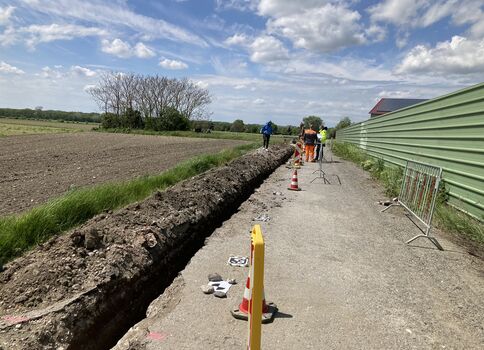 Eine Baustelle an einem Weg: Ein tiefer Graben verläuft links, mehrere Arbeiter in Warnkleidung arbeiten am Rand; Absperrungen, Kegel und Zäune stehen bereit. Rechts eine grüne Metallwand, Felder links, blauer Himmel mit Wolken.