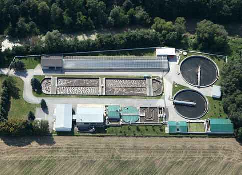 Luftbild eines Kläranlagenkomplexes: mehrere Becken, darunter zwei große runde Reinigungsbehälter, rechteckige Becken, Gebäude und Technik. Die Anlage liegt zwischen Wald und Feldern und dient der Abwasserreinigung.