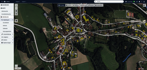 Luftbild einer Satellitenansicht eines kleinen Dorfes mit verwinkelten Straßen und dicht bebauten Häusern in der Ortsmitte; umliegende Felder, Wiesen und Wald umgeben das Siedlungsgebiet.