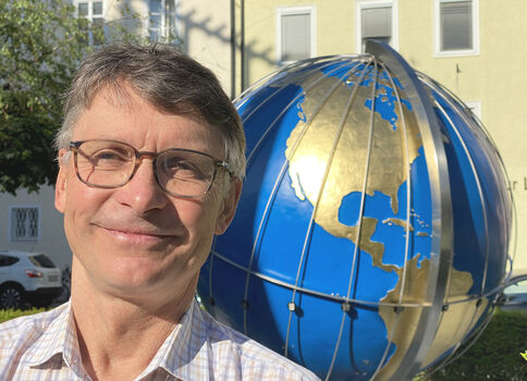 Ein lächelnder Mann mit Brille posiert draußen vor einer großen blauen Globus-Skulptur aus Metall, die goldene Kontinente zeigt; im Hintergrund Häuser einer Stadt.