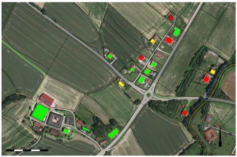 Luftaufnahme eines ländlichen Gebietes mit Feldern und einer Straßenkreuzung. Entlang der Straße stehen mehrere Gebäude, teils mit farbigen Markierungen in Grün, Gelb oder Rot, vermutlich Kennzeichnungen für Status oder Kategorien.