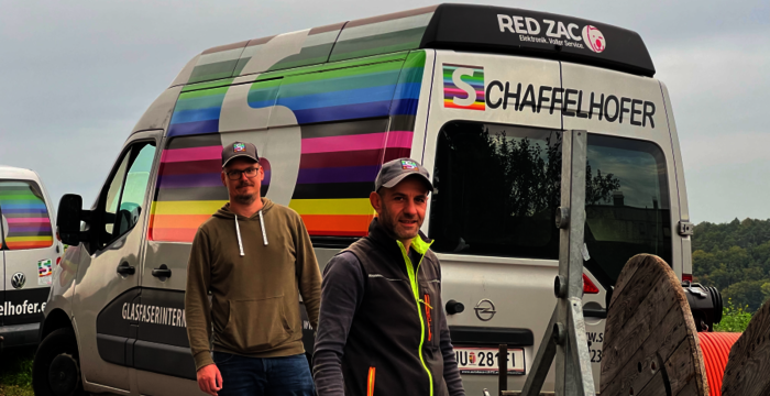 Zwei Männer stehen vor einem silbernen Lieferwagen mit bunten Regenbogenstreifen und Firmenlogos. Das Fahrzeug wirkt wie Werbung; im Hintergrund eine grüne Landschaft.