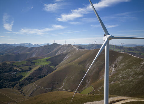 Weite Berglandschaft mit Windkraftanlagen. Ein großes Windrad steht im Vordergrund, weitere Türme finden sich auf den Hügeln im Hintergrund. Blaues Himmelsband, grün-braunes Gelände und kurvige Straßen durch das Tal.