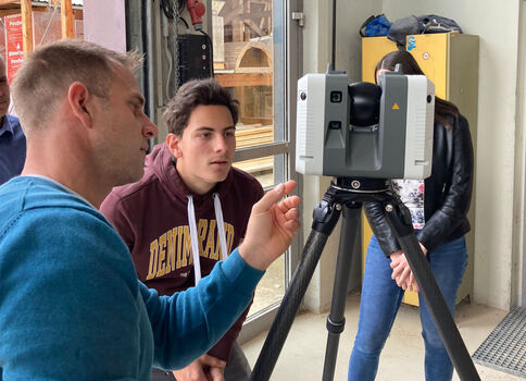 Ein Mann in blauem Pullover erklärt ein Vermessungsgerät auf einem Stativ, während eine Gruppe ihm aufmerksam zuschaut; im Hintergrund befindet sich eine Werkstatt.