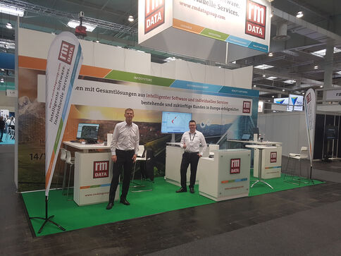Ausstellungsstand von RM Data auf einer Messe. Zwei Mitarbeiter in Hemden stehen an weißen Theken; im Hintergrund Banner, Infostände und eine grüne Teppichfläche. Der Stand präsentiert Software- und Servicelösungen für IT.