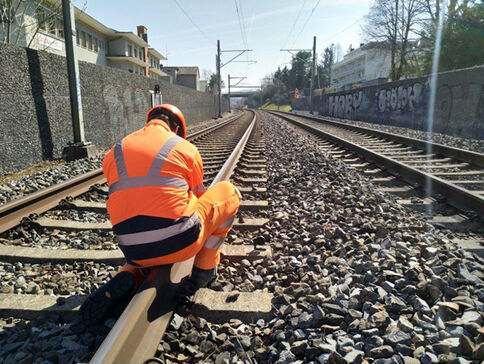 Ein Gleisarbeiter in leuchtend orange Warnkleidung kniet auf einem Bahngleis und führt Wartungsarbeiten aus; im Hintergrund verlaufen weitere Gleise, eine Graffiti-Wand rechts und Häuser links, bei hellem Tageslicht.