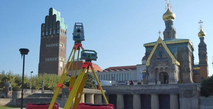 Vordergrund: Vermessungsgerät auf einem Dreibein-Stativ. Hintergrund: orthodoxe Kirche mit goldenen Kuppeln vor klarem blauen Himmel.