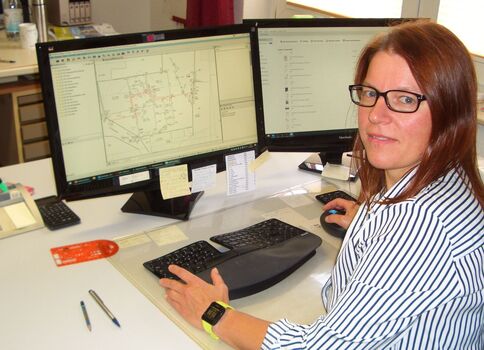 Eine Frau mit Brille sitzt an einem Büro-Schreibtisch vor zwei Monitoren. Der linke Bildschirm zeigt eine Kartendarstellung/Diagramm, der rechte weitere Softwarefenster. Sie tippt auf der Tastatur und trägt ein gestreiftes Hemd.