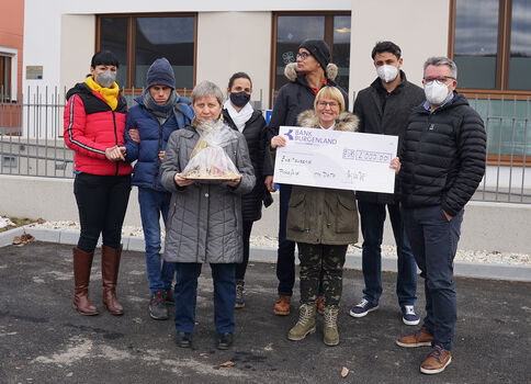 Gruppe von neun Personen steht vor einem modernen Gebäude. Eine ältere Frau in grauem Mantel hält einen transparenten Geschenkkorb; daneben präsentiert eine andere Person einen großen Scheck (Bank Burgenland). Die Szene wirkt wie eine Spendenübergabe.