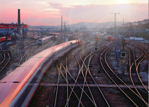 Langzeitbelichtete Aufnahme eines Bahnbetriebs: zahlreiche Gleise kreuzen ein Bahngelände, während links ein schneller Zug eine rote Lichtspur hinterlässt. Im Hintergrund Industrieanlagen und eine warme Abendsonne am Himmel.