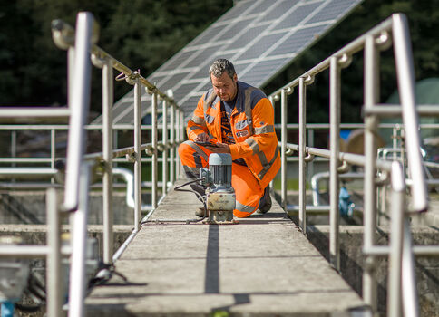 Ein Bauarbeiter in leuchtend orange Warnkleidung kniet auf einer schmalen Brücke mit Geländern und bedient ein handgehaltenes Werkzeug auf einer Betonfläche, vermutlich Wartungs- oder Bauarbeiten.