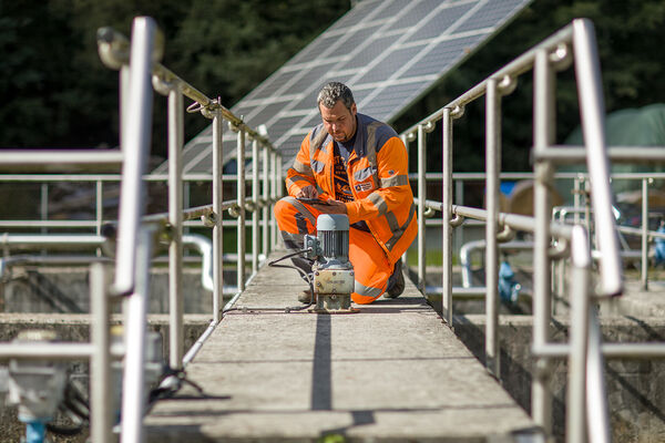 Ein Bauarbeiter in leuchtend orange Warnkleidung kniet auf einer schmalen Brücke mit Geländern und bedient ein handgehaltenes Werkzeug auf einer Betonfläche, vermutlich Wartungs- oder Bauarbeiten.