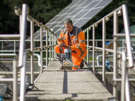 Ein Bauarbeiter in leuchtend orange Warnkleidung kniet auf einer schmalen Brücke mit Geländern und bedient ein handgehaltenes Werkzeug auf einer Betonfläche, vermutlich Wartungs- oder Bauarbeiten.