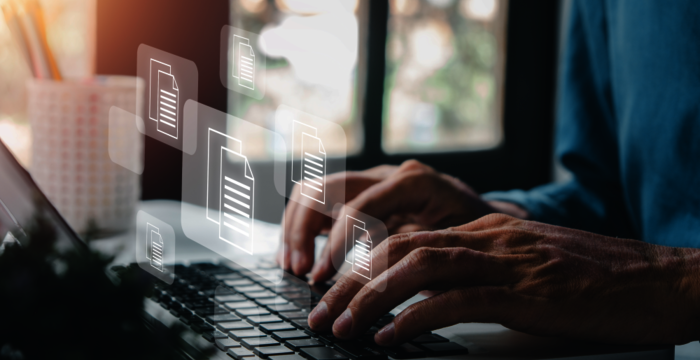 Nahaufnahme von Händen, die auf einer Laptop-Tastatur tippen; darüber schweben transparente Dokumente, die digitale Arbeiten symbolisieren. Hintergrund unscharf mit warmem Fensterlicht.