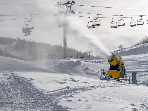 Eine gelbe Schneekanone erzeugt Schnee auf einer Skipiste. Im Hintergrund fährt ein Sessellift über die verschneite Berglandschaft; der Himmel ist bedeckt.