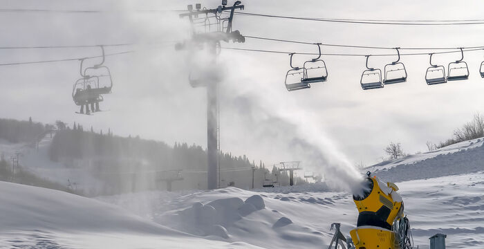 Eine gelbe Schneekanone erzeugt Schnee auf einer Skipiste. Im Hintergrund fährt ein Sessellift über die verschneite Berglandschaft; der Himmel ist bedeckt.