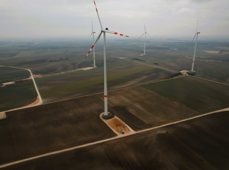 Luftbild eines Windparks: Mehrere Windkraftanlagen mit rot-weißen Rotorblättern stehen auf flachen, braunen Feldern; eine Straßenführung verbindet die Turbinen, im Hintergrund erstrecken sich weitere Windräder am Horizont.