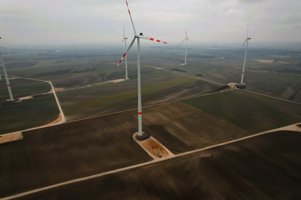 Luftbild eines Windparks: Mehrere Windkraftanlagen mit rot-weißen Rotorblättern stehen auf flachen, braunen Feldern; eine Straßenführung verbindet die Turbinen, im Hintergrund erstrecken sich weitere Windräder am Horizont.