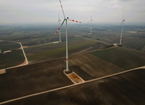 Luftbild eines Windparks: Mehrere Windkraftanlagen mit rot-weißen Rotorblättern stehen auf flachen, braunen Feldern; eine Straßenführung verbindet die Turbinen, im Hintergrund erstrecken sich weitere Windräder am Horizont.