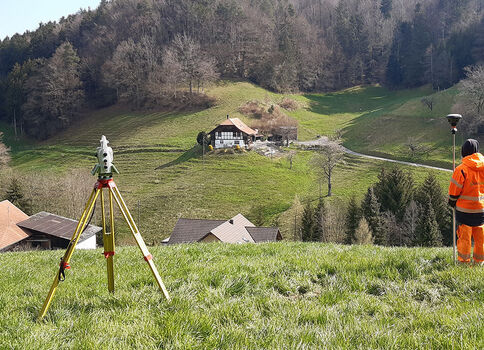 Un technicien en tenue haute visibilité orange se tient dans un pré vert, près d’un trépied de relevé topographique. À l’arrière-plan, collines boisées et quelques maisons blanches dans une vallée paisible.