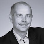 Schwarz-weißes Porträt eines freundlich lächelnden Mannes in Sakko und Hemd, frontal zur Kamera aufgenommen.