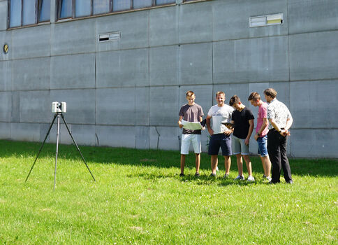 Fünf junge Männer stehen im Gras vor einer grauen Gebäudewand und schauen auf Unterlagen, während ein Kameratripod mit Kamera aufgebaut ist.